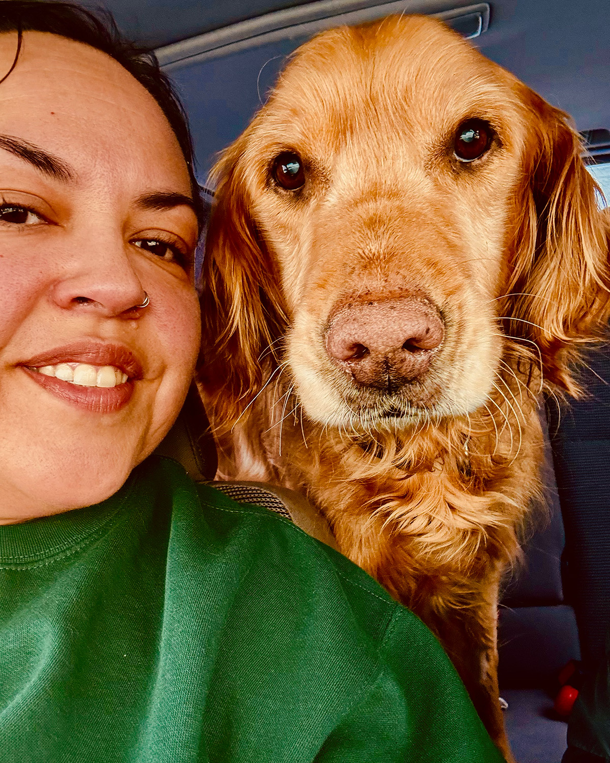 Melanie smiling in the front seat of a car next to Deuce, a golden retriever with soulful eyes, both looking into the camera.
